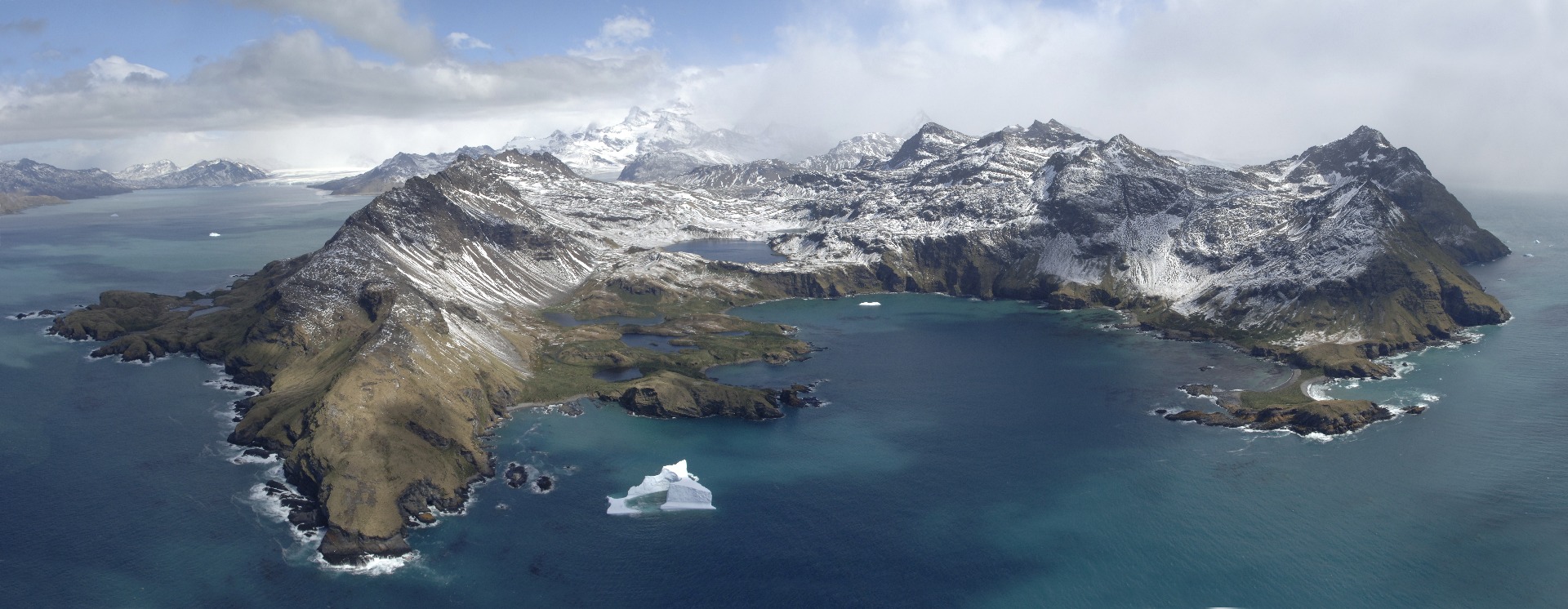 Oblique aerial image of South Georgia; a snow-dusted mountainous island in the sea, with an iceberg